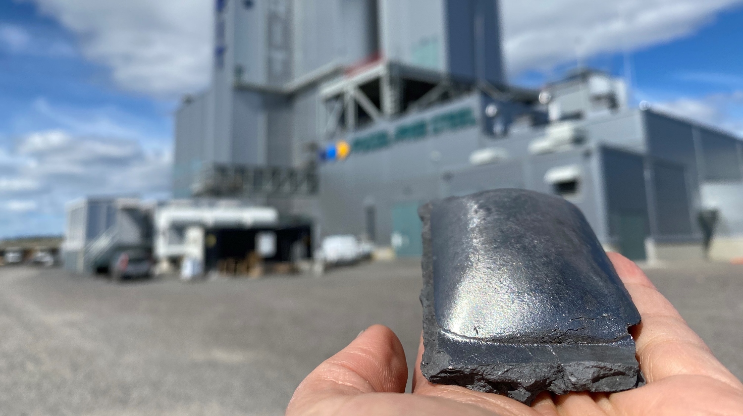 An image of a someone holding a piece of sponge iron wit the Hybrit fossil-free steel plant in the background.