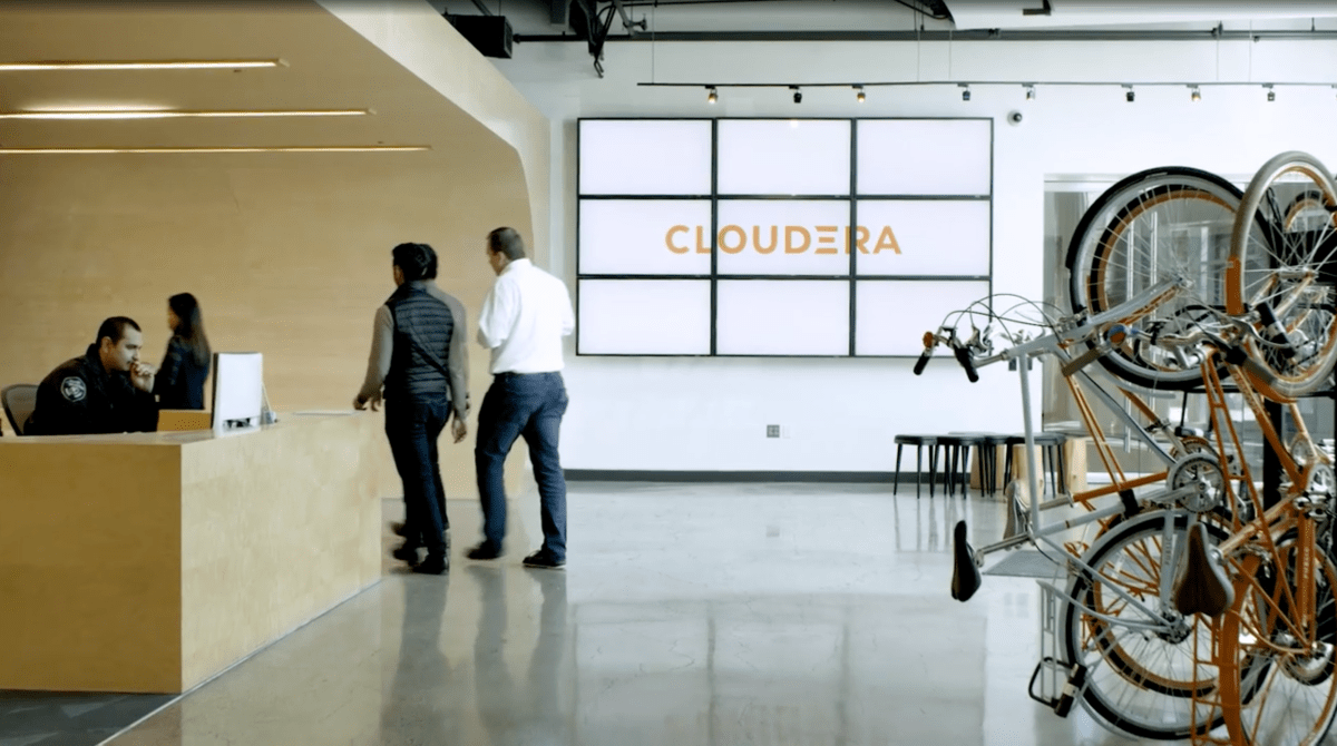 Cloudera office with guard behind wooden desk on left, bikes in standing bike rack on the right, and several people passing a large Cloudera logo in the foreground.