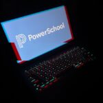 a laptop in a dark room with the laptop keyboard lit up with a red and blue glitchy effect, and the display reads PowerSchool with its corporate logo.