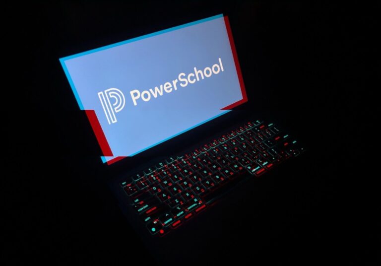 a laptop in a dark room with the laptop keyboard lit up with a red and blue glitchy effect, and the display reads PowerSchool with its corporate logo.