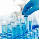 A close-up shot of a laboratory technician's gloved hand holding a test tube with a blue liquid among a rack of other test tubes.