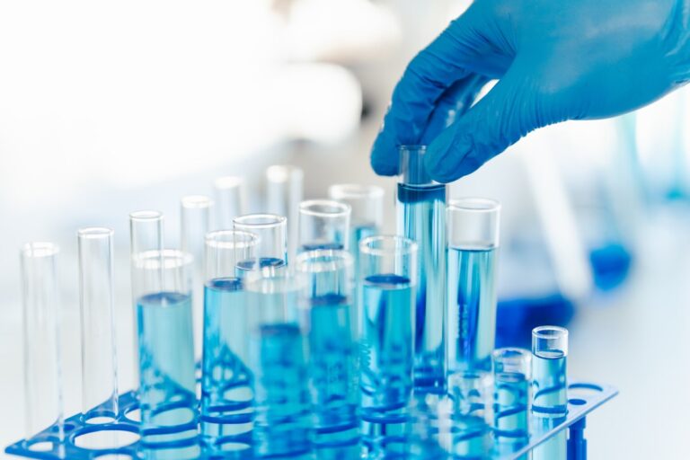 A close-up shot of a laboratory technician's gloved hand holding a test tube with a blue liquid among a rack of other test tubes.